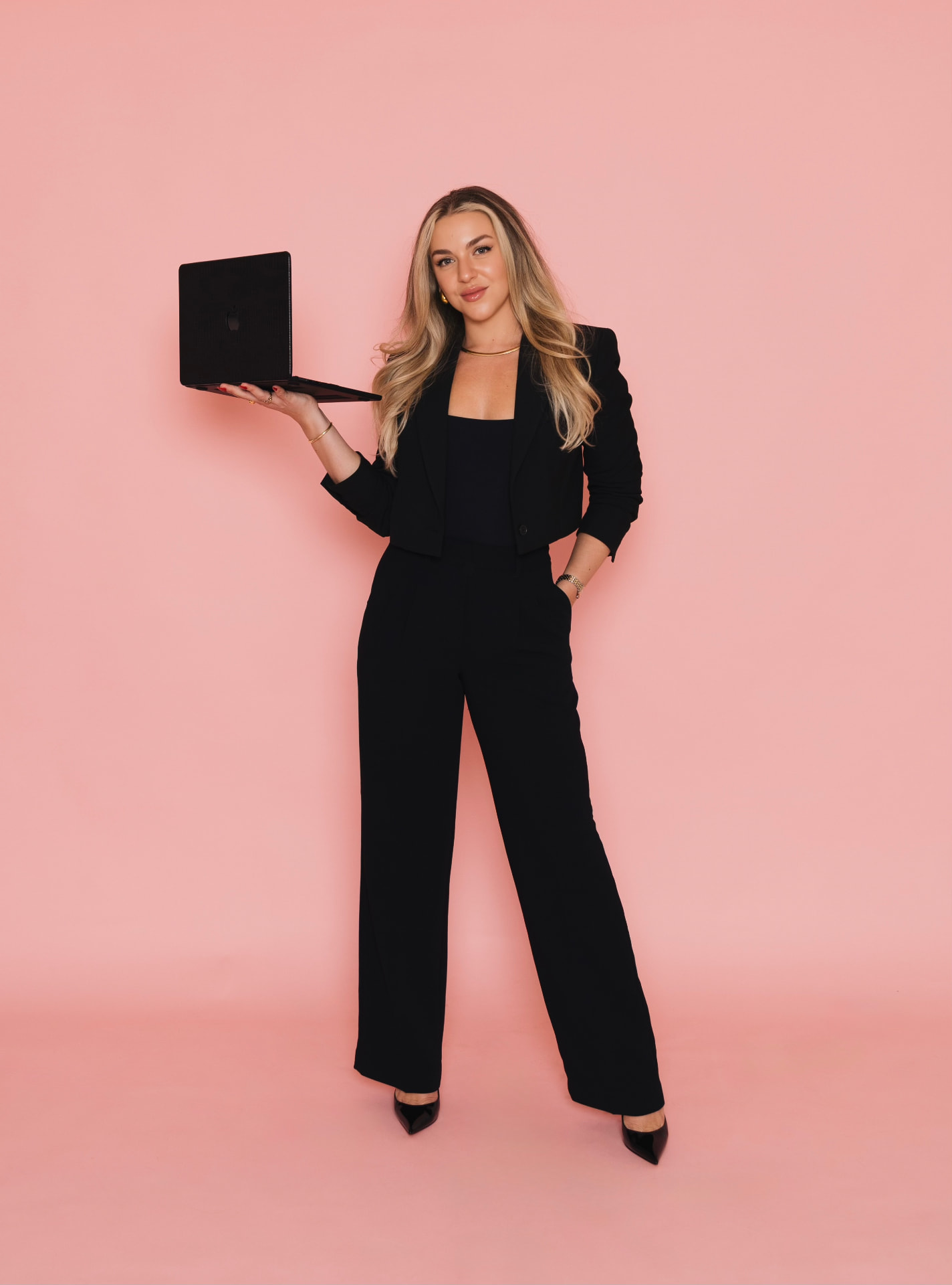 Gina De La Guardia in a black suit holding a laptop against a pink backdrop